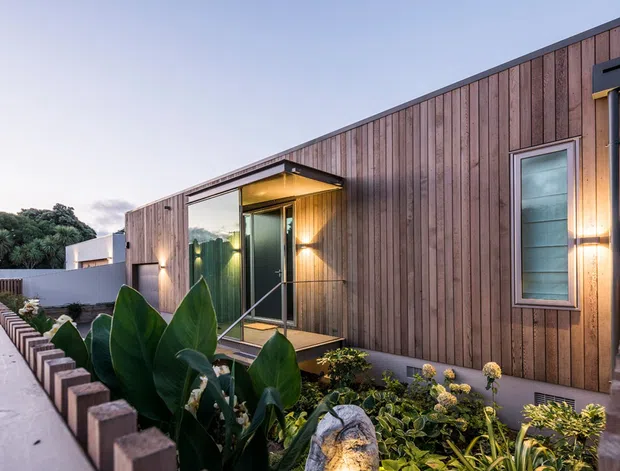 A contemporary single-storey residential facade clad in vertical timber boards, with a recessed glazed entry, narrow horizontal window, integrated exterior lighting, and landscaped planting beds bordered by low brick walls and concrete paths.