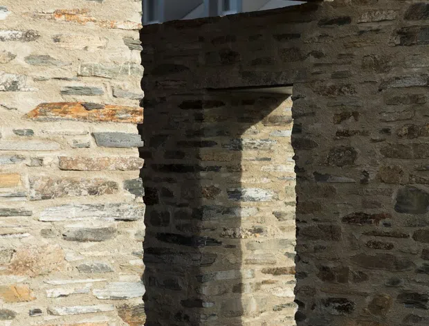 Close architectural view of a rustic stone structure with thick rough masonry walls, narrow openings, and exposed dark timber roof and beam construction. Strong sunlight creates deep shadows across the irregular stone surfaces, emphasizing the material texture and structural layering.