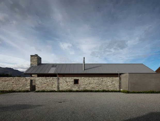 A low, single-storey building viewed from the exterior, featuring a long dark metal pitched roof, rough natural stone masonry walls, a stone chimney, and a plain rendered end wall. The building sits beside a broad gravel-paved foreground, with mountainous terrain visible in the distance under a partly cloudy sky. A small portion of a timber-clad neighbouring structure is visible at the far right.