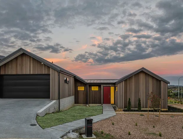 Exterior view of a contemporary single-storey home at dusk, composed of connected gabled volumes clad in vertical timber, with a dark garage door, concrete block base, red entry door, paved walkway, gravel landscaping, and a small landscaped garden. The neighbouring buildings and sunset sky form the background.