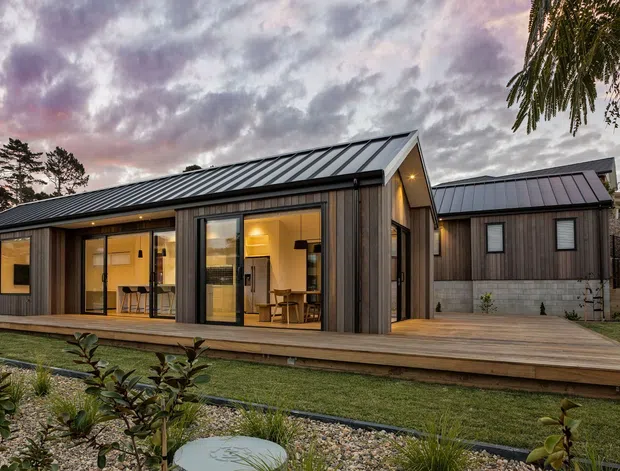 Single-storey contemporary residential building viewed from the landscaped yard at dusk. The home has dark vertical timber cladding, black standing-seam pitched roofs, extensive glazed sliding doors, and a broad timber deck connecting the main pavilion to a smaller adjoining wing. Warm interior lighting is visible through the glazing, with low garden planting and stone landscaping in the foreground.