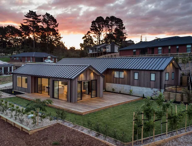 Exterior view of a contemporary single-storey residential building at dusk, featuring dark vertical timber-style cladding, extensive glazing, and a low-pitched corrugated metal roof. The home opens onto a timber deck and landscaped garden with raised planting beds, while similar residences and tall trees form the background.