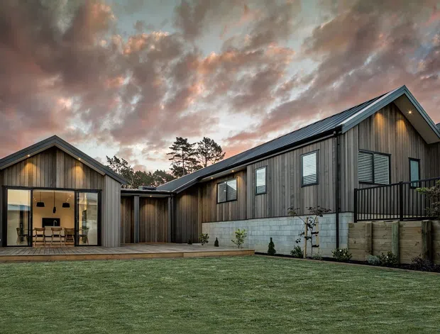 Exterior view of a contemporary single-storey home composed of connected pitched-roof volumes. The building features dark vertical timber cladding, a light concrete-block base, large glazed openings, timber decking, and landscaped lawn, photographed at dusk beneath a dramatic cloudy sky.
