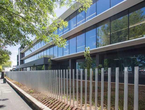 A contemporary multi-storey commercial building viewed from a tree-lined sidewalk. The facade features extensive dark-framed floor-to-ceiling glazing, horizontal projecting slab bands, dark cladding, and a vertical metal security fence along a landscaped frontage.
