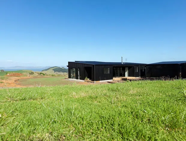 A low, single-storey contemporary house set in an open rural landscape. The building has dark black cladding, a series of connected rectangular volumes with shallow flat or low-pitched rooflines, large windows and glazed openings, and a small timber-toned deck or entry platform. A broad expanse of green grass dominates the foreground, with rolling farmland and distant hills beyond under a clear blue sky.