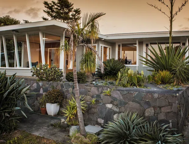 Single-storey modernist house with broad low-pitched roof planes, extensive glazing, slender white structural posts, and warm timber-lined interiors visible through the windows. The home is set behind a raised dark irregular-stone retaining wall and dense drought-tolerant, subtropical planting, including palms, agaves, and succulents.