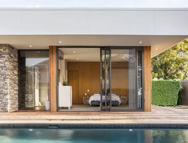 Contemporary single-storey residential facade viewed from a swimming pool. The home features a broad flat roof with a deep covered terrace, floor-to-ceiling glazing and dark framed sliding doors, warm vertical timber cladding around the entry, and a stacked natural-stone column. A bedroom is visible behind the glazing, opening onto a timber deck beside the pool and landscaped hedge.