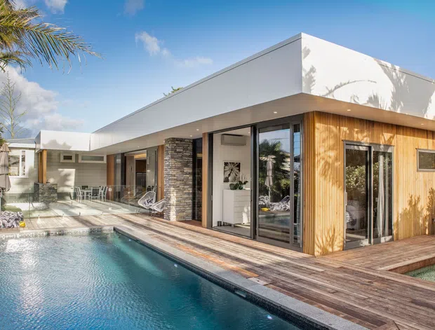 Contemporary single-storey residence with a flat roof, white rendered volumes, warm vertical timber cladding, dark stone feature walls, and extensive glazing. The home opens onto a timber pool deck and a long swimming pool, with a bright tropical outdoor setting.