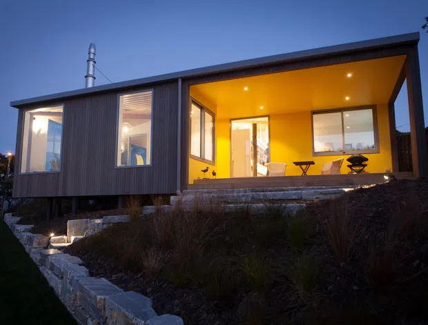 Contemporary single-storey modular-looking house photographed at dusk, with a dramatic cantilevered flat roof and a warm, fully glazed recessed outdoor living area. Dark vertical cladding wraps the enclosed volume, while exposed timber-toned interior ceilings and illuminated glazing create a strong contrast. Stone retaining walls and landscaped terraces sit in front of the elevated home.