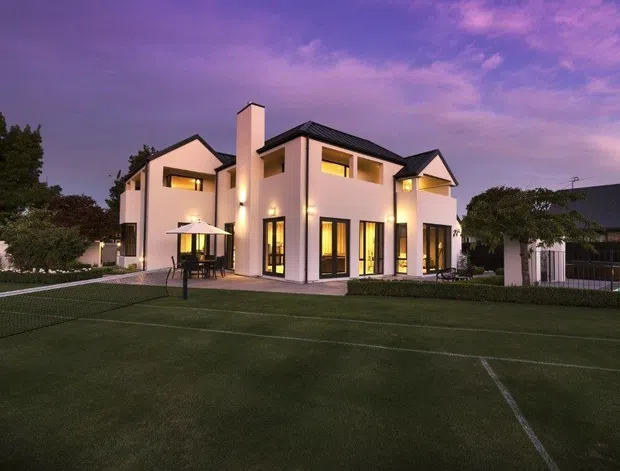 A large contemporary multi-storey residence photographed at dusk, with pale rendered walls, dark pitched roof sections, prominent chimney, tall illuminated windows, and a covered outdoor entertaining area. The property fronts a landscaped lawn containing a marked tennis court, with mature trees and neighbouring structures in the background.