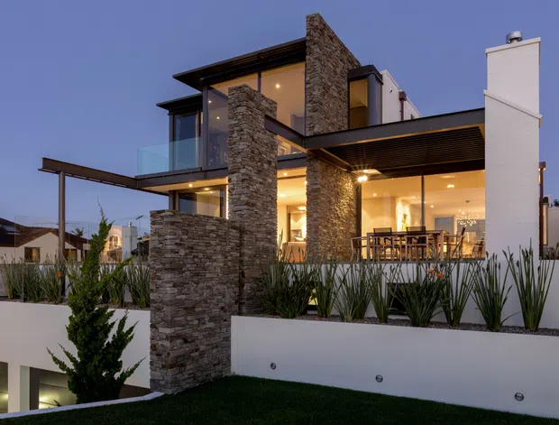 Dusk exterior view of a sophisticated multi-level contemporary residence. The home features extensive floor-to-ceiling glazing, projecting flat roof planes, cantilevered balconies, a covered outdoor dining area, tall white rendered walls and prominent stacked-stone feature columns. Layered planter beds with architectural landscaping frame the foreground.