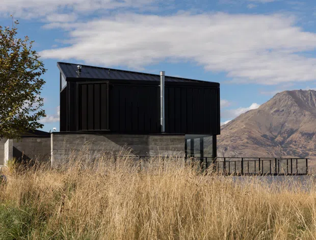 Exterior view of a compact, dark-clad modern building set in a dry grassy landscape, with a low concrete retaining wall, black metal railing and deck or terrace edge. A rugged mountain rises prominently in the background beneath a partly cloudy sky. The building has a simple rectilinear form, a shallow sloping metal roof, vertical dark cladding and large glazed openings.