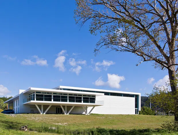 A low, modernist two-storey house set in a landscaped suburban or semi-rural site. The building has a long horizontal composition, broad glazed upper-level facade, white horizontal cladding, slim roof planes, and distinctive angled structural supports beneath the upper floor. A large mature tree and open lawn frame the exterior.