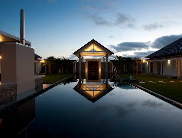 Dusk exterior view of a low-rise architectural complex arranged around a long reflective water feature. A lit, gabled pavilion with strong symmetrical columns forms the focal point, flanked by dark pitched-roof buildings and landscaped courtyards. The scene has dramatic sky reflections, warm architectural lighting, and a refined resort-like atmosphere.