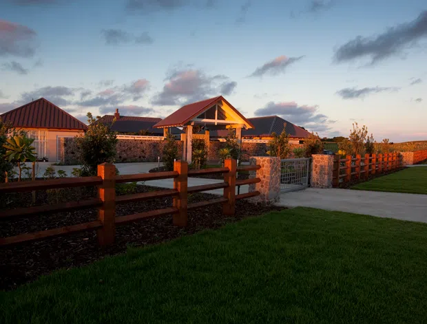 A single-storey rural-style property viewed from a landscaped exterior at dusk. The complex has multiple pitched and gabled roofs, warm timber framing, stone masonry walls, metal roofing, and a covered central entry or pavilion. Timber post-and-rail fencing, a paved driveway or walkway, lawn, and open agricultural land frame the building.