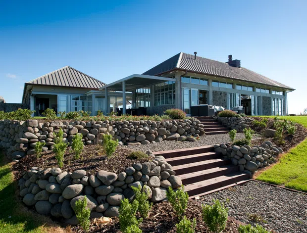 Exterior view of a low-rise residential home set on a landscaped sloping site. The house has pale weatherboard-style walls, extensive glazed openings, a covered outdoor connection or veranda, and pitched corrugated metal roofs. Broad timber steps lead through rounded natural-stone retaining walls and garden beds to the lawn.