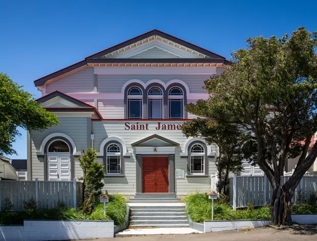 Front-on exterior view of a historic church-like building with a symmetrical classical facade, prominent triangular pediment, decorative gable detailing, arched windows, horizontal weatherboard cladding, and a central red entrance door. The building is painted pale grey and pink with dark red trim, set behind low fencing and mature trees under a clear blue sky.