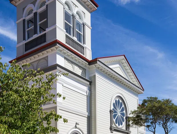 Exterior view of a tall, ornate white weatherboard building with a prominent tower, red metal roof, decorative cornices, arched fanlight windows, classical mouldings, and dark balustrades. The facade has a formal heritage character, with a driveway and parked cars at ground level and a bright blue sky behind.