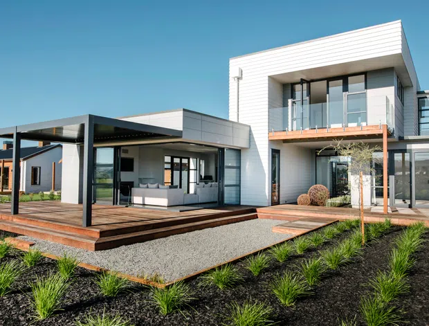 Contemporary two-storey residence with a crisp white and charcoal facade, large glazed openings, a flat-roofed projecting carport or covered outdoor structure, and expansive timber decking. The landscaped front garden uses dark mulch, gravel paths, structured grasses, and clipped shrubs. A balcony with glass balustrading sits above the recessed entry, while a furnished living area is visible through the glazing.