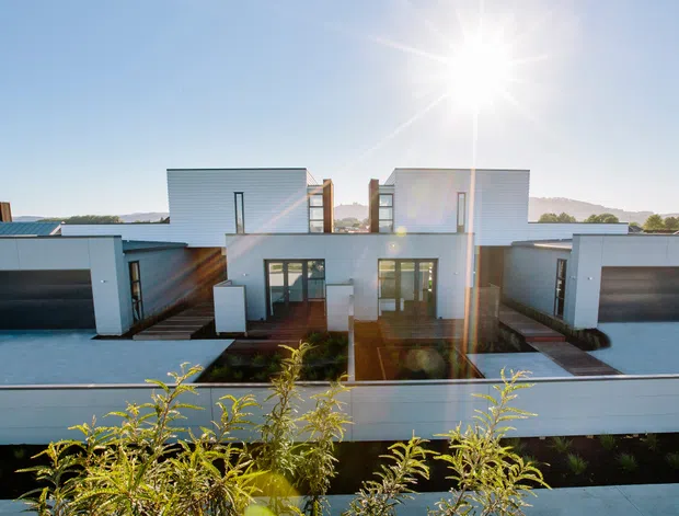Contemporary multi-level residential facade viewed from the street, featuring crisp white rendered volumes, dark timber or metal accents, large glazed entry doors, integrated double garages, and a low horizontal boundary wall. A distant waterfront or landscape setting is visible beyond the property.