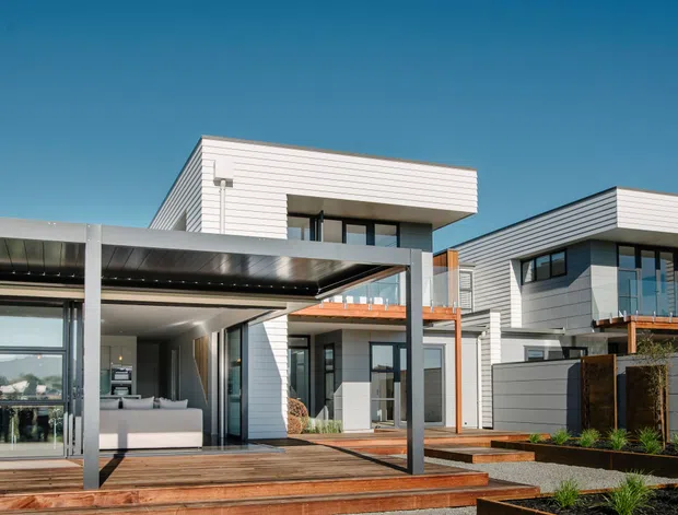 Exterior view of a modern multi-level residential home with crisp white horizontal cladding, dark-framed glazing, projecting flat roof forms, and a large covered outdoor entertaining deck. The façade combines white and charcoal-grey panels with timber decking and warm timber-toned balcony accents, set against a clear blue sky.