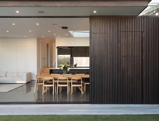 A contemporary open-plan home viewed from an outdoor terrace, showing a dining area and kitchen behind full-height glazing, with a living room to the left. The facade includes a prominent vertical dark timber screen, while the interior features pale walls, timber dining furniture, dark kitchen cabinetry, polished dark flooring, recessed lighting, and a skylight.
