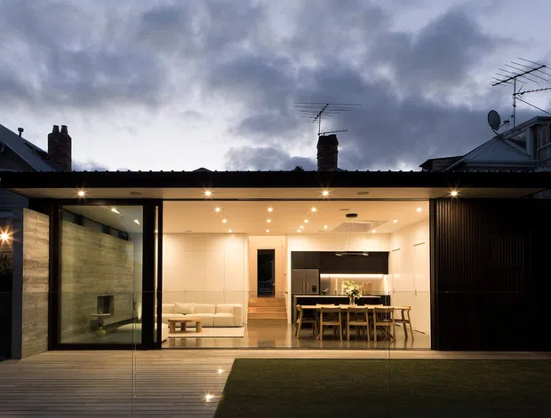 Evening exterior view through full-height glazing of a contemporary rear extension, showing an open-plan living and dining area with a modern kitchen. The low-profile addition has a broad flat roof, black-framed glazing, dark vertical cladding, a timber deck, and warm recessed interior lighting. Older neighbouring rooflines and chimneys are visible behind the extension.