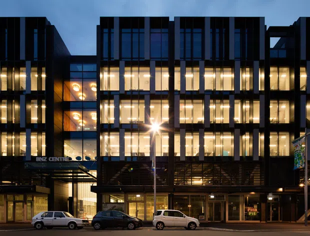 Evening exterior view of a multi-storey contemporary commercial building with a dark, strongly gridded facade and extensive illuminated glazing. The street-level frontage includes retail or lobby spaces, with cars parked along the road and signage visible on the building.
