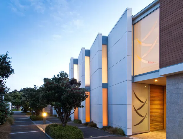 Twilight exterior view of a contemporary multi-unit residential facade along a landscaped pedestrian walkway. The building features tall projecting vertical fins clad in smooth light-grey panels, warm illuminated recesses, dark horizontal wood-look cladding, textured masonry, large decorative glazed entry panels, and timber-toned entry doors. Low hedges, trees, and pathway lighting frame the frontage.