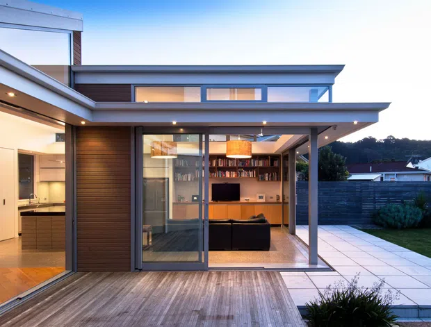 Contemporary multi-level home with a strong indoor-outdoor connection, viewed from a timber deck and paved terrace. The open living room features extensive floor-to-ceiling glazing, built-in timber shelving and cabinetry, dark-framed doors, and warm pendant lighting. Flat roof planes, projecting eaves, and a mix of dark horizontal timber cladding and smooth light walls define the modern facade. A paved outdoor terrace and landscaped garden are visible to the right.