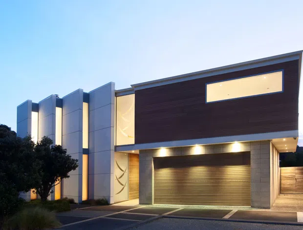 Contemporary multi-level residential facade photographed at dusk, featuring strong geometric vertical wall panels, illuminated recessed glazing, a dark horizontal timber-look upper facade, a wide roller garage door, and a sheltered driveway entry. Landscaping and neighboring elements are secondary.