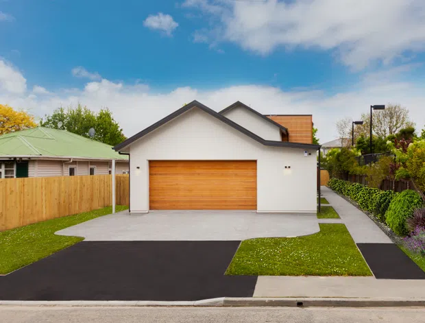 Street-facing view of a contemporary suburban house facade dominated by a detached-looking double garage with a white rendered exterior, broad timber garage door, dark pitched roof, concrete forecourt, black asphalt driveway, and a paved pedestrian path. A low green-roofed neighbouring structure, timber fencing, hedges, and landscaped planting are visible around the property.