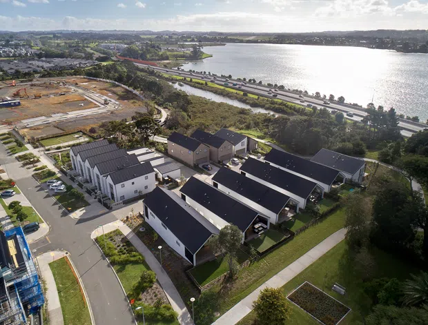 Aerial view of a recently developed residential neighbourhood beside a broad waterway, with rows of detached and semi-detached homes, dark pitched roofs, landscaped gardens, internal streets and footpaths. A major road runs along the waterfront, with urban development and low hills visible in the distance.