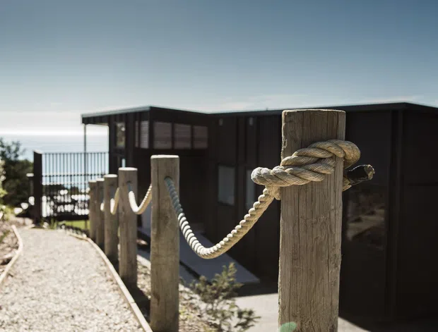 Exterior view of a dark, modern flat-roofed building beside a gravel walkway, with substantial timber posts connected by thick nautical rope forming a balustrade or boundary. A railing and bright water-like horizon are visible beyond the building, while sparse coastal vegetation frames the scene.
