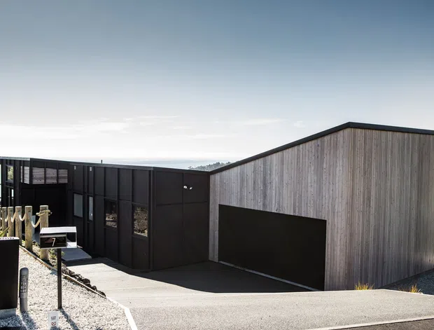 Exterior view of a contemporary single-storey building on a gently sloping site. The composition features a dark, metal-clad wing with extensive glazing and a larger volume clad in vertical weathered timber, with a broad dark garage door and paved driveway in the foreground. Sparse landscaping and open rural surroundings are visible beyond the building.