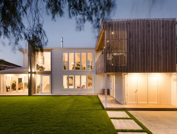 Dusk exterior view of a contemporary multi-level residence with a white rendered or clad lower volume, extensive vertical glazing, a projecting upper volume screened in closely spaced vertical timber battens, and an integrated double garage. A manicured lawn and rectangular stepping-stone path lead toward the entry and garage terrace, while warm interior and exterior lighting emphasises the facade.