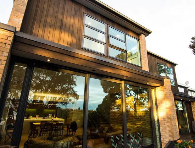 Two-storey contemporary residential extension or house facade with warm brown brick masonry, vertical timber wall cladding, dark-framed floor-to-ceiling glazing, and a projecting upper level. The glazing reveals a dining and living area inside, while a paved outdoor area and landscaping are visible around the building.