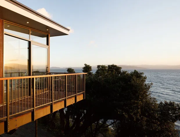 Exterior view of a contemporary elevated coastal building with a projecting timber-clad balcony, slender metal balustrade, broad glazing, and a flat roof. The structure overlooks a large body of water with distant hills visible across the horizon, while mature vegetation partially screens the lower level.