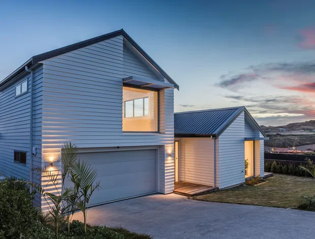 Exterior view of a contemporary two-storey house at dusk, clad in pale horizontal weatherboards with dark metal pitched roofs. The facade includes an integrated double garage, illuminated upper-level glazing, a recessed entry porch, driveway, and landscaped planting. A hilly suburban setting is visible beyond the property.