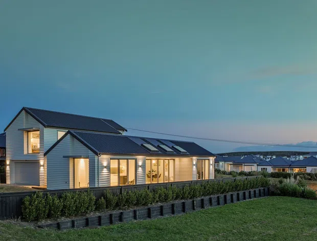 Exterior twilight view of a contemporary single-storey residential house with layered pitched gable roofs, dark metal roofing, light horizontal cladding, warm illuminated windows, and landscaped hedges. Similar homes extend into the background across a broad suburban development, with distant hills visible on the horizon.