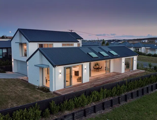 Exterior dusk view of a contemporary single-storey residential home with white horizontal weatherboard cladding, dark pitched metal roofs, multiple skylights, and extensive glazed doors opening onto a timber deck. The house sits in a landscaped suburban development with low retaining walls and neighboring homes visible in the background.