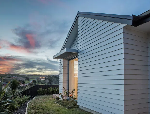 Exterior view of a modern single-storey home at dusk, featuring crisp white horizontal weatherboard cladding, a sharply pitched gable end, dark metal roof and trim, and a sheltered entry canopy. The house sits on a landscaped sloping suburban site with low planting and distant hills beneath a dramatic evening sky.