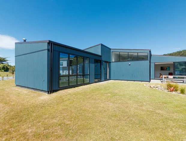 Exterior view of a modern single-storey building composed of several interconnected cubic volumes. The facade is clad in dark blue-grey vertical ribbed metal panels, with extensive black-framed glazing and a partially covered outdoor dining or seating area. The building sits within a broad lawn and landscaped garden under a clear blue sky, with a low wooded backdrop.