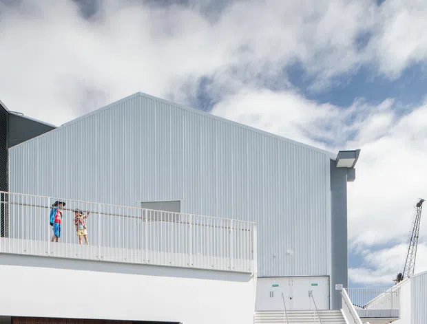Exterior view of a contemporary multi-level building with a large corrugated light-grey metal-clad gable volume above a white lower level. A broad external stair and upper balcony with white metal railings dominate the facade, while a dark timber-clad entrance sits below. Bicycles and people are present in the foreground, with a construction crane visible at right beneath a bright, partly cloudy sky.