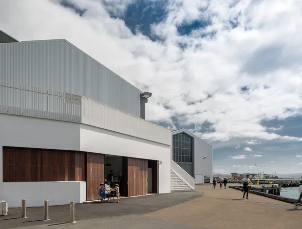 Exterior view of a contemporary waterfront commercial or civic building with corrugated light-grey metal cladding, white rendered walls, large dark timber entrance doors, and a prominent glazed gable-like end wall. The building opens onto a broad paved promenade beside a marina or harbour, with a cloudy coastal sky and distant waterfront structures visible.