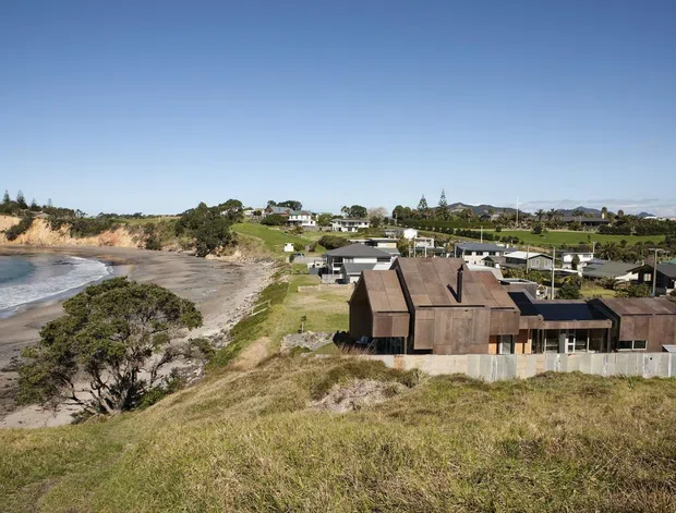 Aerial view of a coastal residential property on a grassy, elevated site overlooking a sandy beach and bay. The main home has a low-profile, multi-gabled form with dark timber-clad walls and roof elements, extensive glazing, and a pale boundary fence. Additional suburban homes and vegetation are visible inland.