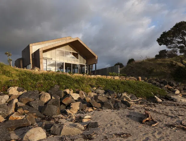 A modern angular house set on a rocky coastal or waterfront site, viewed from the shoreline. The building has a strong pitched or gabled roof form, extensive floor-to-ceiling glazing on the front elevation, dark vertical cladding, and a warm timber-toned roof or fascia. Native-looking vegetation, exposed rocks, and sandy shoreline surround the structure beneath a dramatic cloudy sky.