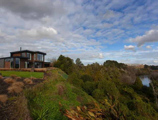 A modern multi-level house with extensive floor-to-ceiling glazing sits on a landscaped, elevated site overlooking dense native vegetation and a small body of water. The home features dark cladding, timber-toned framing, flat rooflines, and a projecting upper level beneath a dramatic partly cloudy sky.