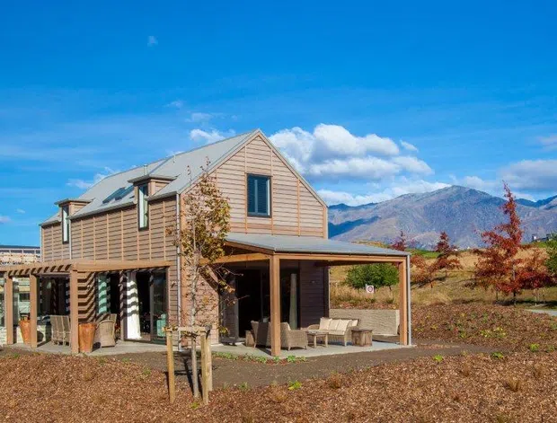 Exterior view of a contemporary two-storey timber-clad house with a steep gabled roof, covered outdoor terrace, pergola, and extensive glazing. The home sits in a landscaped rural or semi-rural setting with mountains visible in the background.