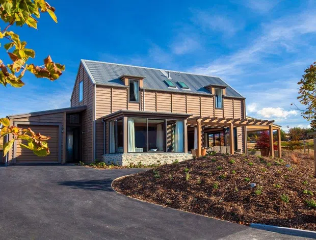 Exterior view of a two-storey timber-clad house with a pitched corrugated metal roof, dormer-style roof windows, large glazed living-area windows, an attached garage, and a timber pergola extending over the outdoor area. The house sits beside a newly landscaped garden and paved driveway under a bright blue sky.